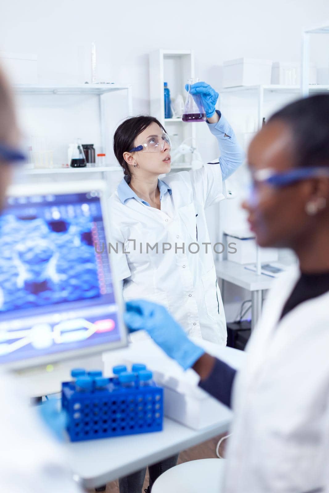 Serious thoughtful scientist analyzing color for liquid in glass flask. Multiethnic team of medical researchers working together in sterile lab wearing protection glasses and gloves.