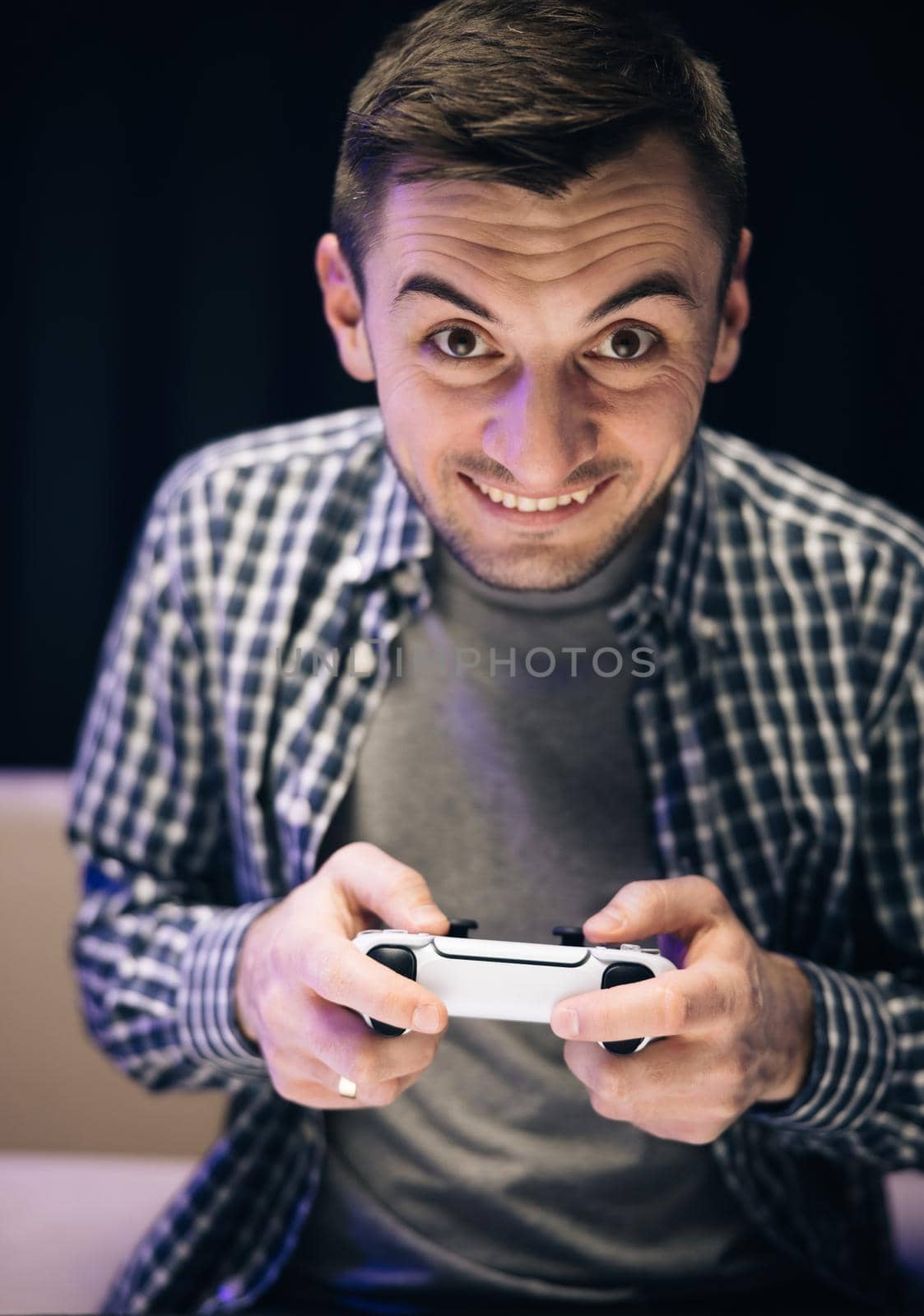 Man in casual wear playing video game in home living room. Caucasian guy holding joystick control having fun laughing on couch indoors by uflypro