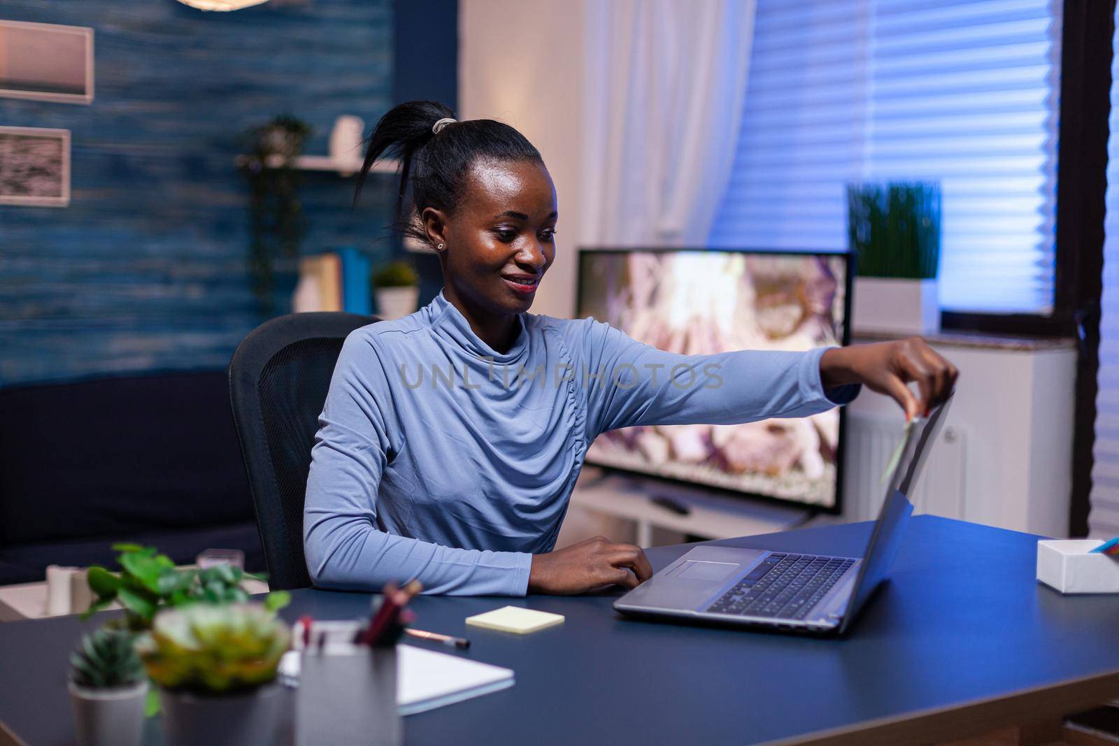 African business woman smiling at webcam during conference call in the evening sitting at desk. Black entrepreneur sitting in personal workplace writing on keyboard.