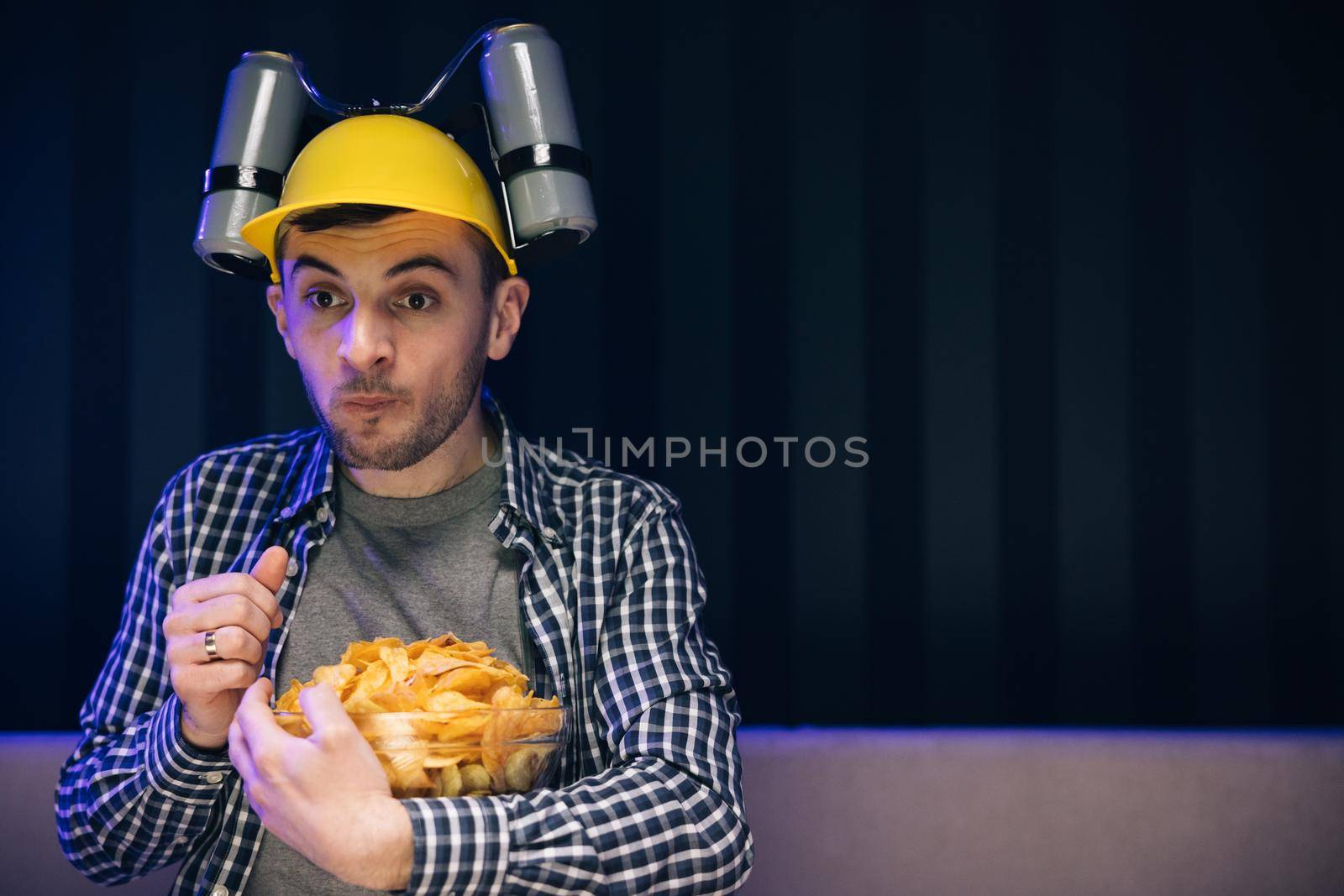 Man with beer helmet on the head eats chips while sitting at home on couch, in evening he watches movies at home on TV. Caucasian man rests at home and watches TV shows or sports news on TV screen by uflypro