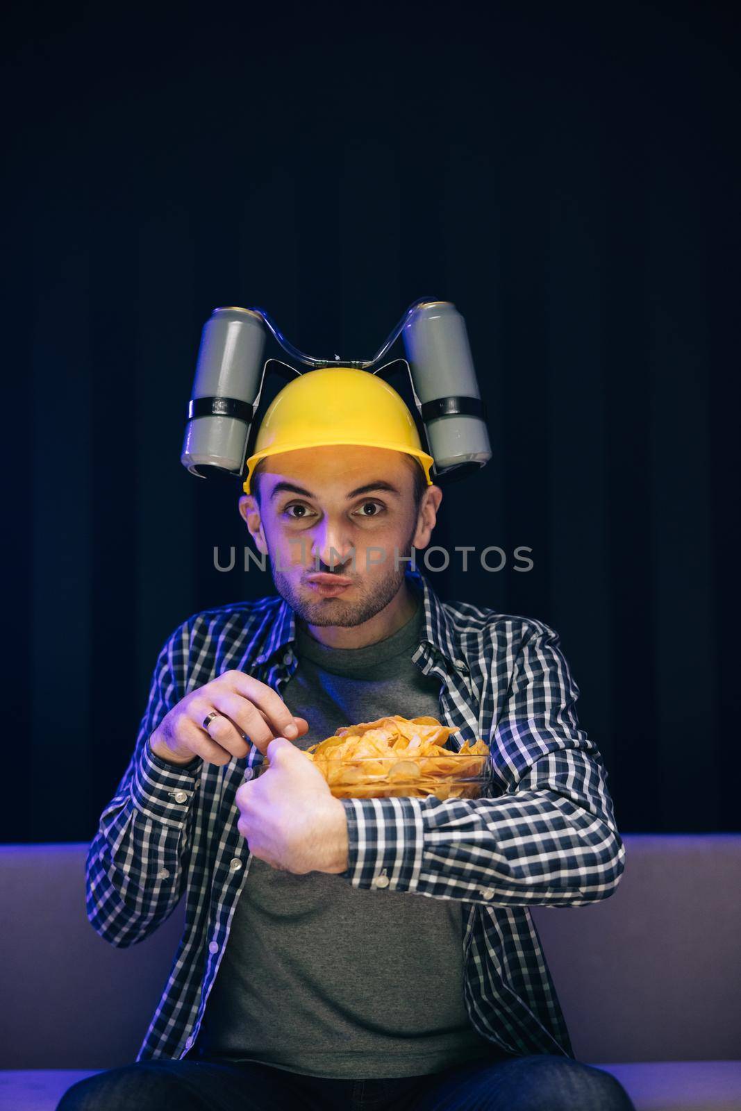 Young funny man with beer helmet on the head eating chips while watching movie on the sofa in the living room.