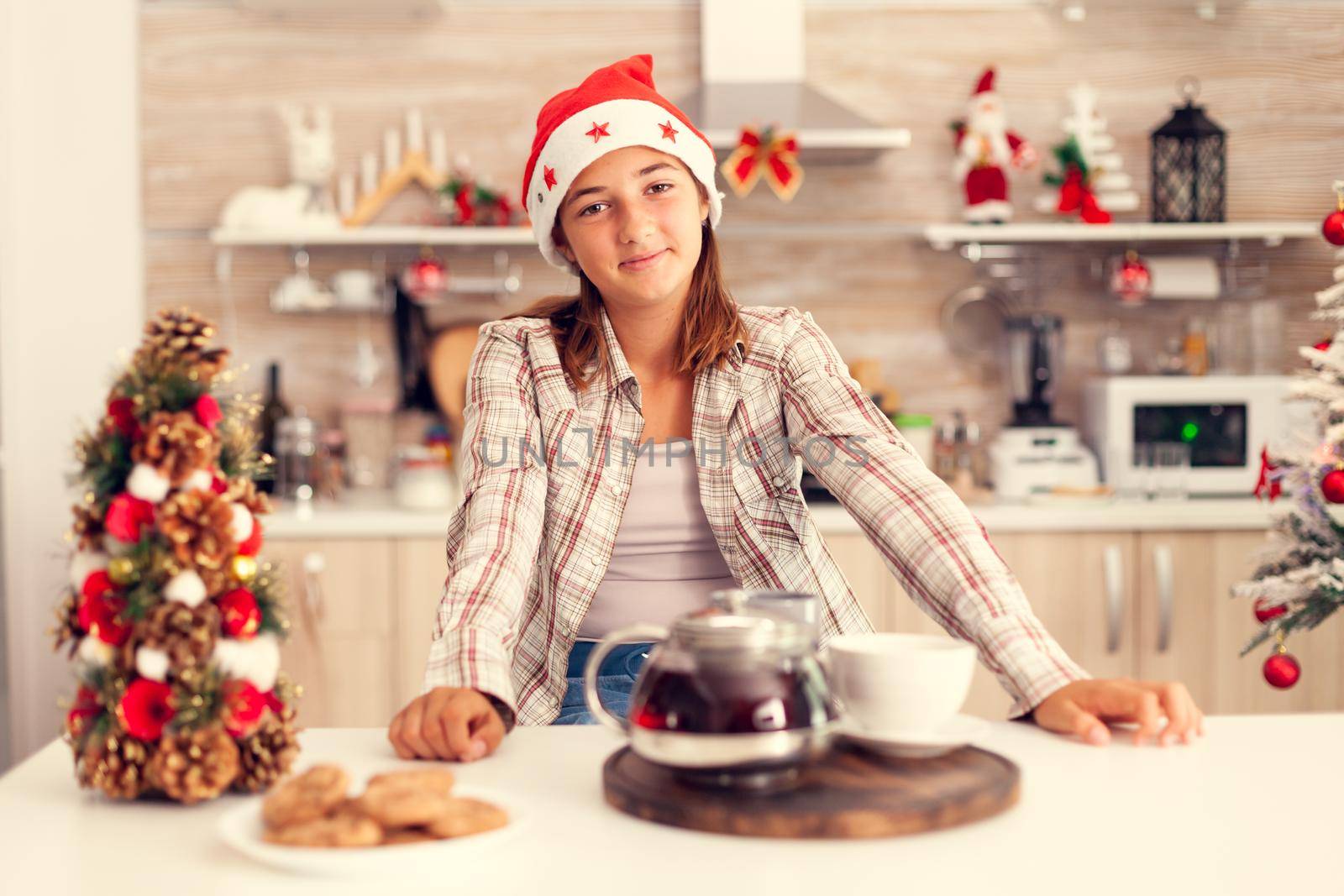 Joyful girl wearing santa heat celebrating christmas in kitchen. Cheerful happy adorable teenager girl in home kitchen with delicious biscuits and xmas tree in the background celebrating winter holiday.