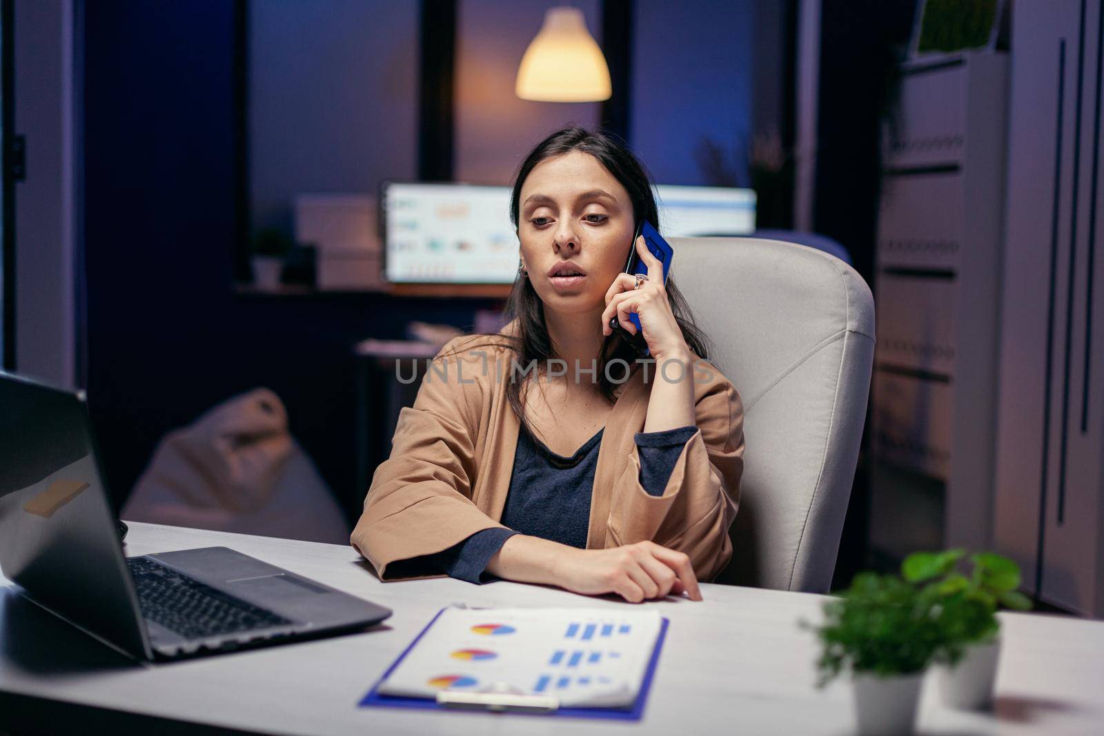 Workaholic manager talking with customer over the phone in the evening. Woman entrepreneur working late at night in corporate business doing overtime in the course of phone call.