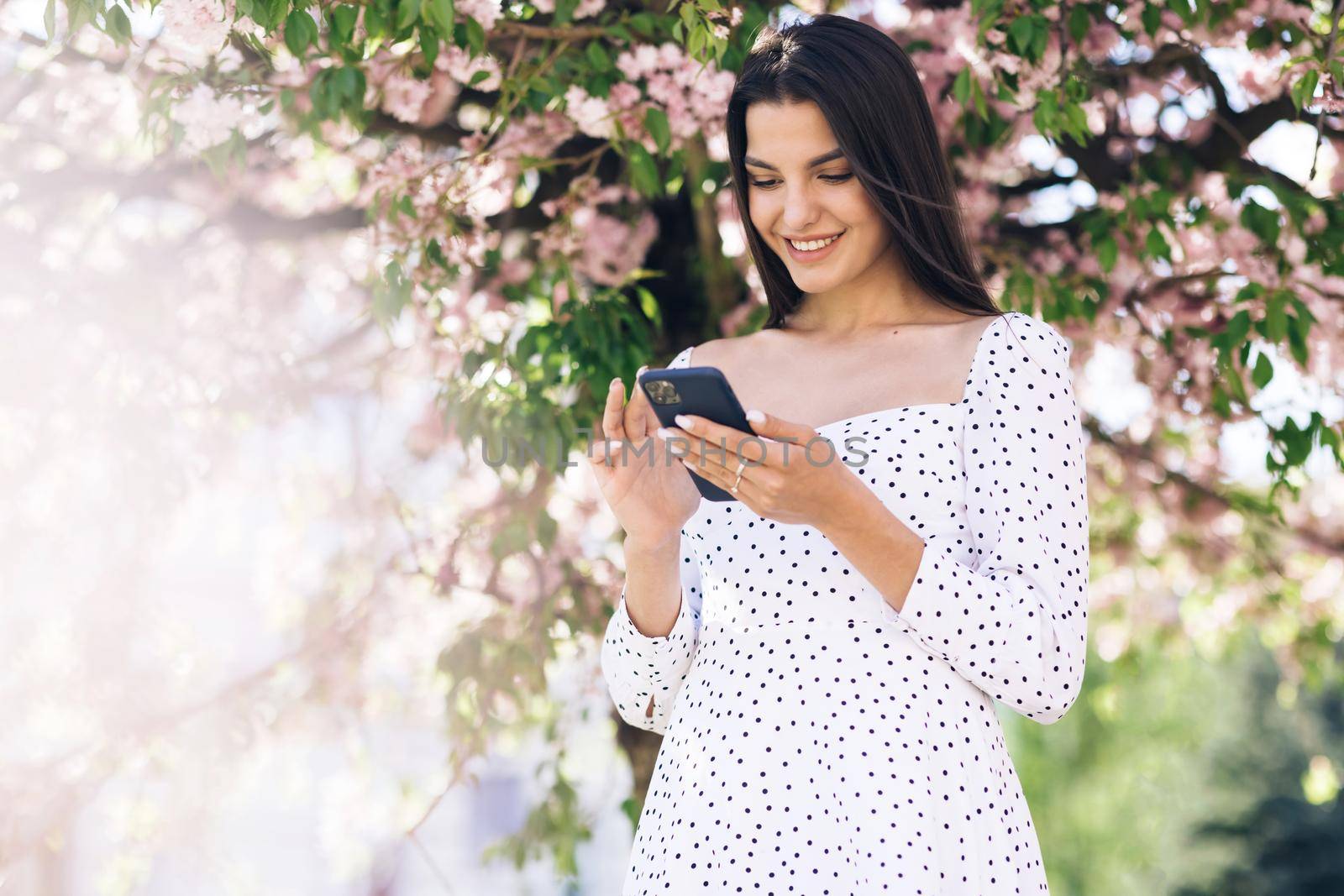 Smiling woman wearing white dress using smartphone. Girl using social media application text messages receive news smiling outdoor. Communication, social networks, online shopping concept. Technology by uflypro