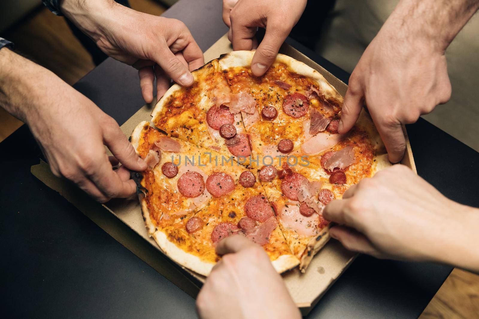 Male hands taking slices of pizza with cheese, tomatoes and ham from food delivery. Group of hungry friends sitting at desk and sharing delicious lunch on table background by uflypro