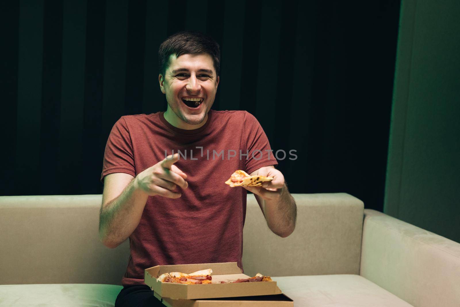Portrait of young hipster eating pizza, watching favorite comedy on tv while relaxing at sofa in the living room. Happy man eating Italian food. by uflypro