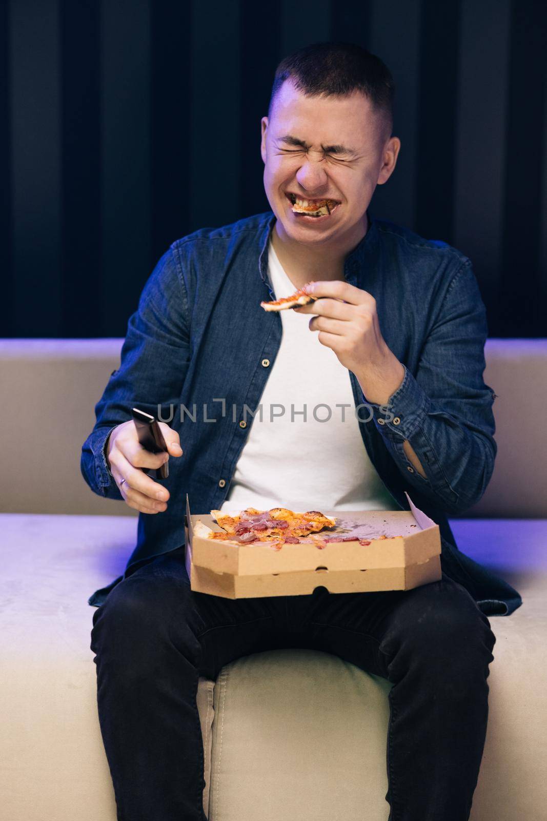 Man eating pizza sitting on sofa smiling at home. Relaxation resting. Funny young emotional caucasian fan man watching a game, eating pizza and reacting to team playing.