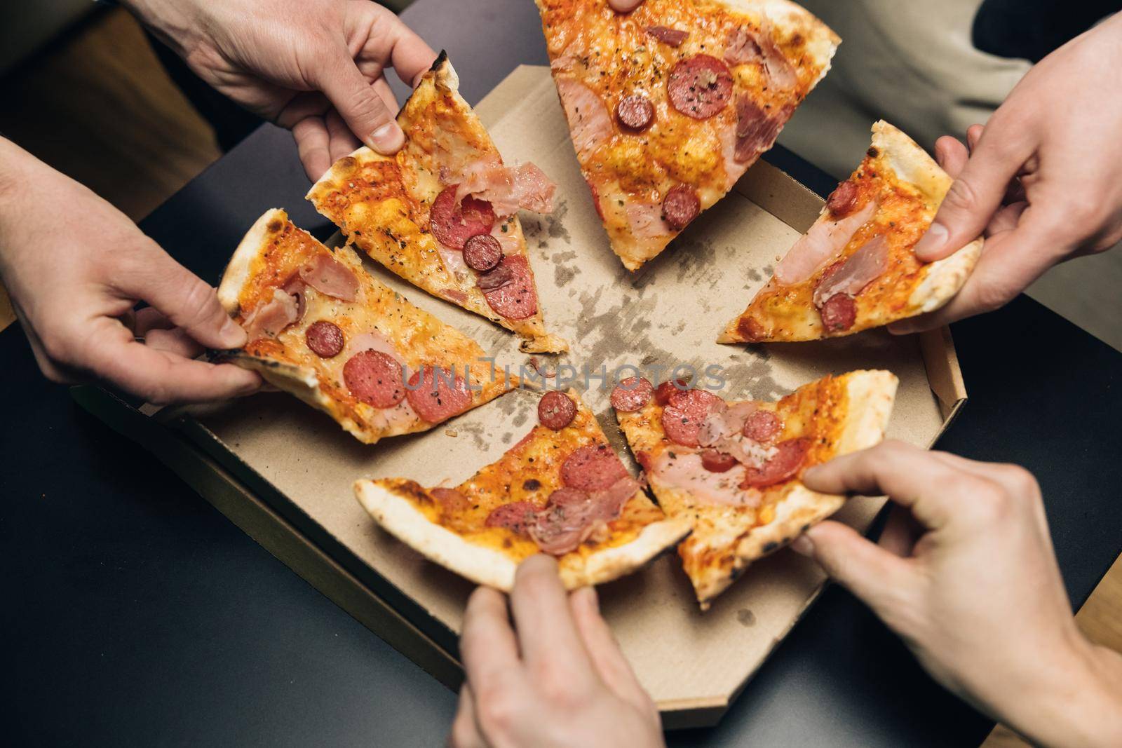 Group of hungry friends sitting at desk and sharing delicious lunch on table background. Male hands taking slices of pizza with cheese, tomatoes and ham from food delivery by uflypro