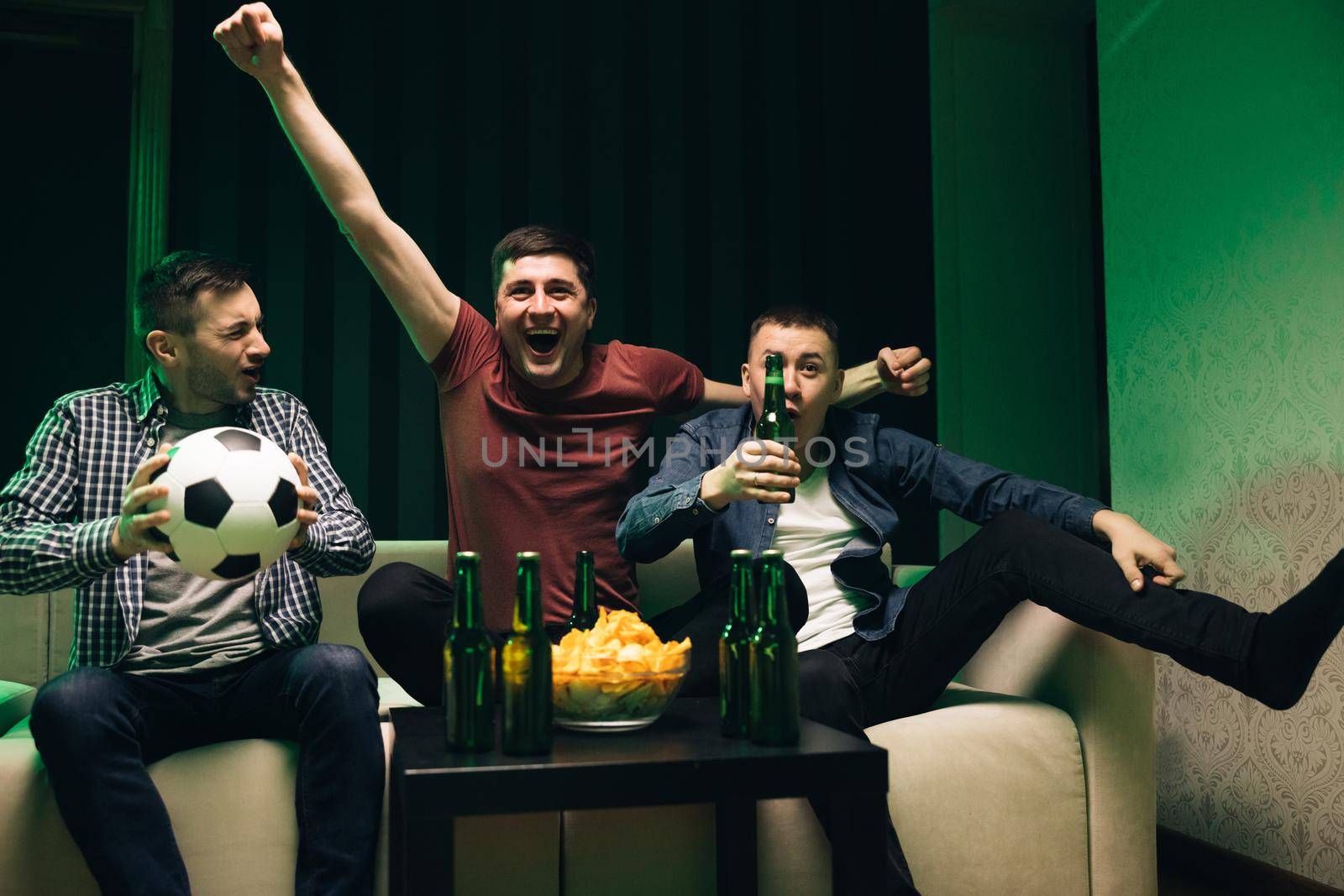 Male friends watching sport channel gathered together. Three handsome happy caucasian friendly men eating chips snacks and cheering up for football game on TV while sitting on sofa.