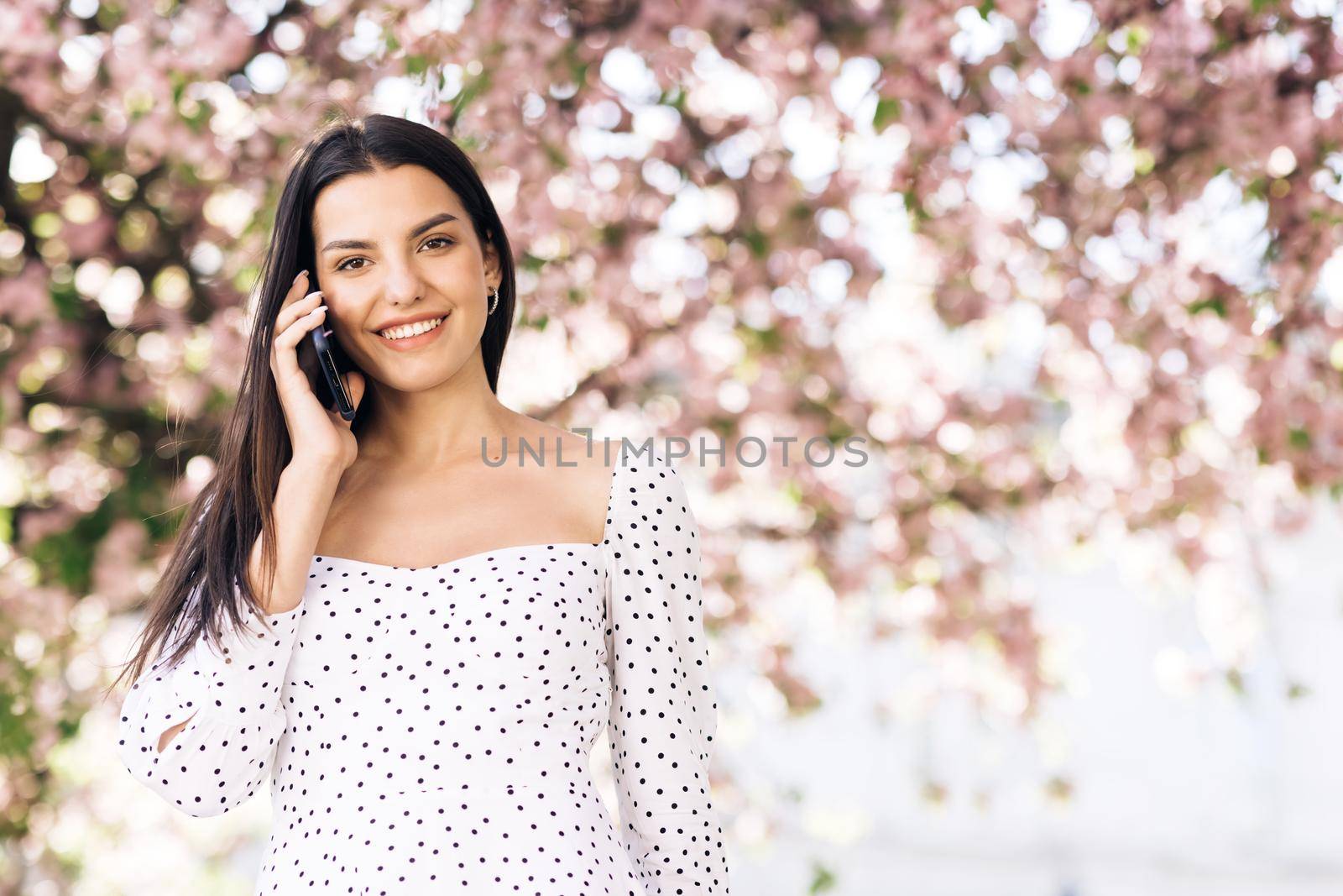 Young model girl dressed in a white summer dress talking on the phone walking in the park on a background of sakura trees. Attractive girl with long hair talks on a smartphone and smiles outdoor by uflypro