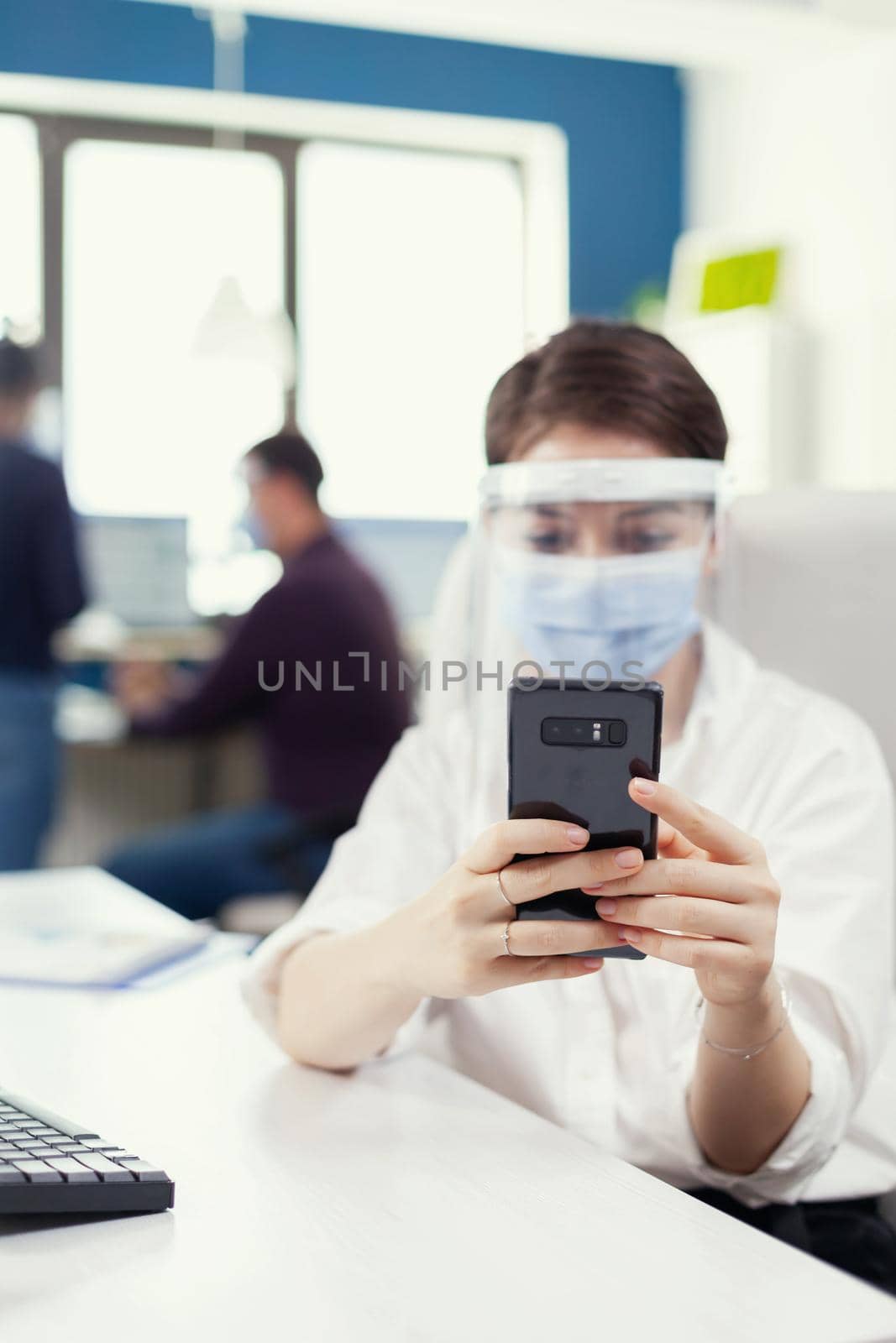 Businesswoman using mobile phone texting on smartphone wearing face mask as safety precaution against covid19. Multiethnic coworkers working respecting social distance in financial company.