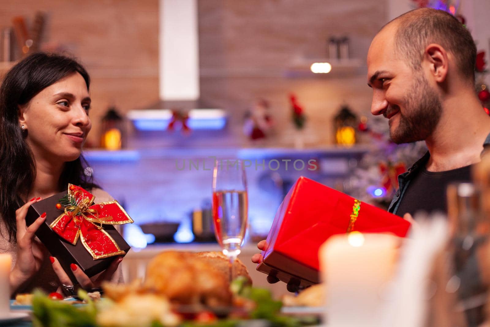 Happy couple surprising each other with present gift with ribbon on it enjoying christmastime together sitting at xmas table in decorated kitchen. Smiling family celebrating christmas season