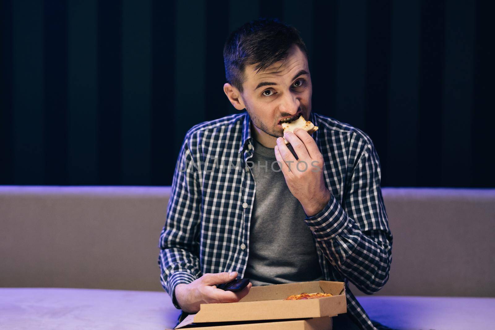 Handsome young man eating pizza while watching tv while relaxing at sofa in the living room by uflypro