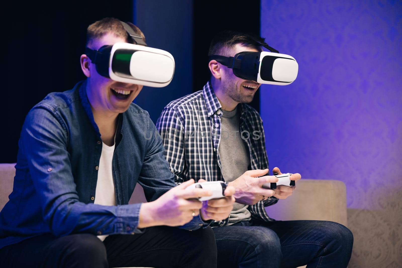 Overjoyed men friends winning videogame having fun together on sofa celebrating victory at home. Excited funny young gamers wear virtual reality glasses holding controllers playing video game by uflypro