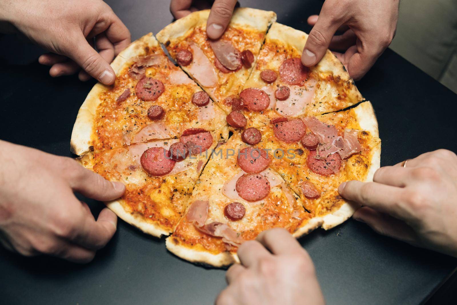 Top view male hands taking slices of pizza with cheese, tomatoes and ham from food delivery. Group of hungry friends sitting at desk and sharing delicious lunch on table background by uflypro