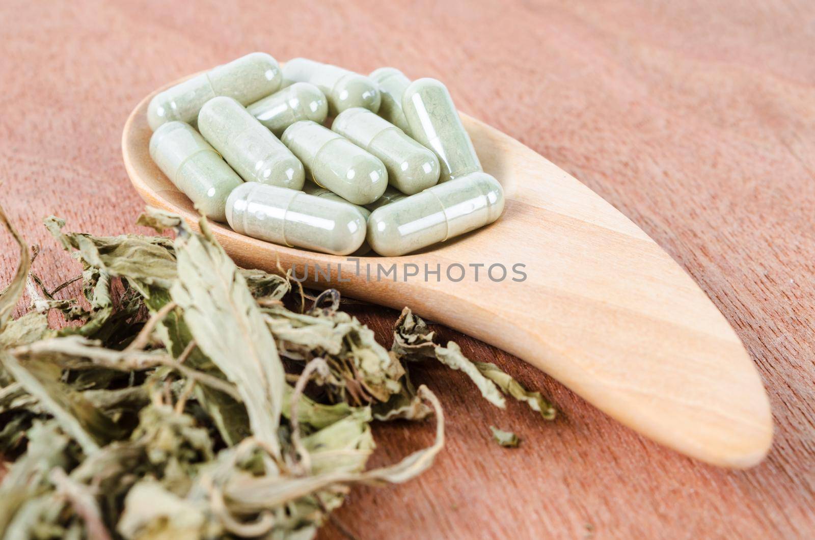Pile of herbal medicine capsules in wooden spoon with dried leaves on wooden background.