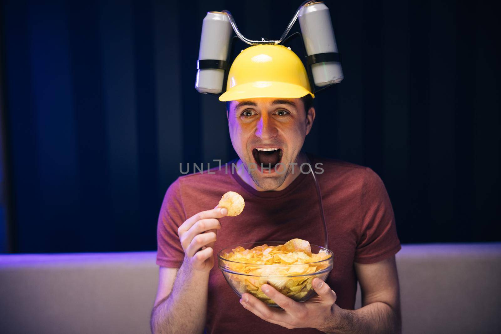 Young funny man with beer helmet on the head eating chips while watching movie on the sofa in the living room by uflypro