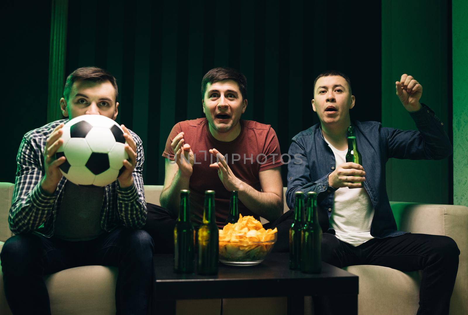 Three handsome happy caucasian friendly men eating chips snacks and cheering up for football game on TV while sitting on sofa. Male friends watching sport channel gathered together