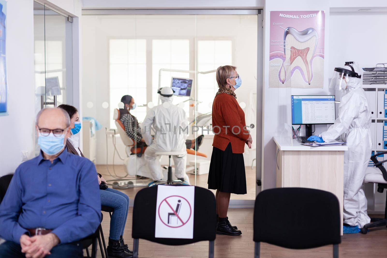 Senior patient with face msak discussing with dentist assistant dressed ppe suit keeping social distancing in waiting area. Dentistiry staff in protection suit during global pandemic with covid-19.