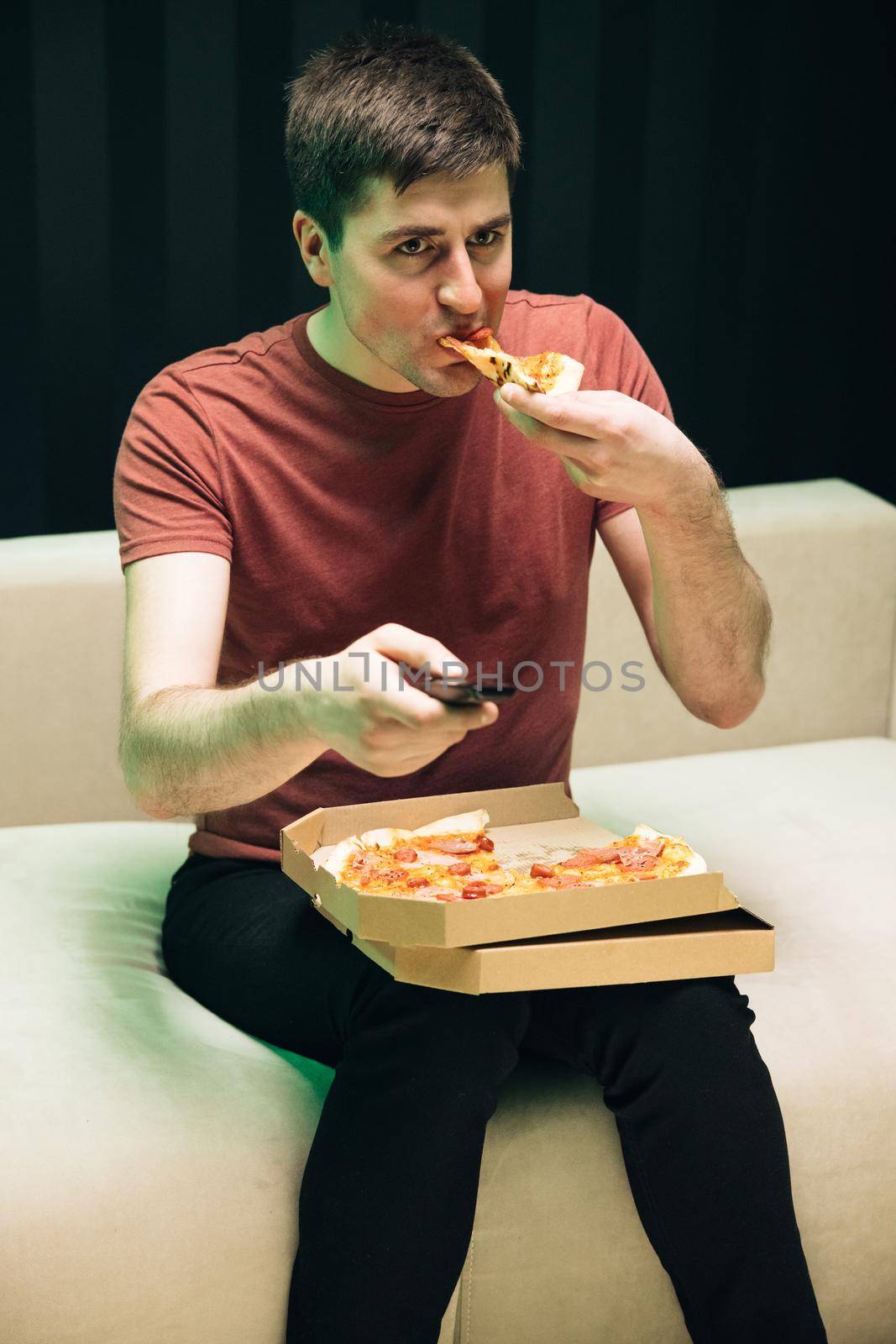 Relaxation cinema channels resting. No emotions, communication problem. Portrait of young man watching TV eating pizza sitting on sofa at home.