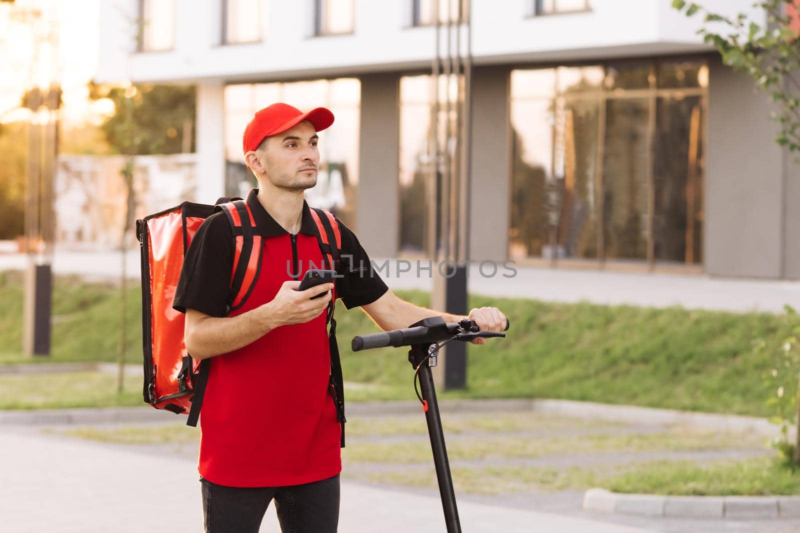 Male courier food delivery with red thermal backpack walks street with electric scooter uses smartphone navigate. Deliveryman worker employee deliver online order client customer at sunset