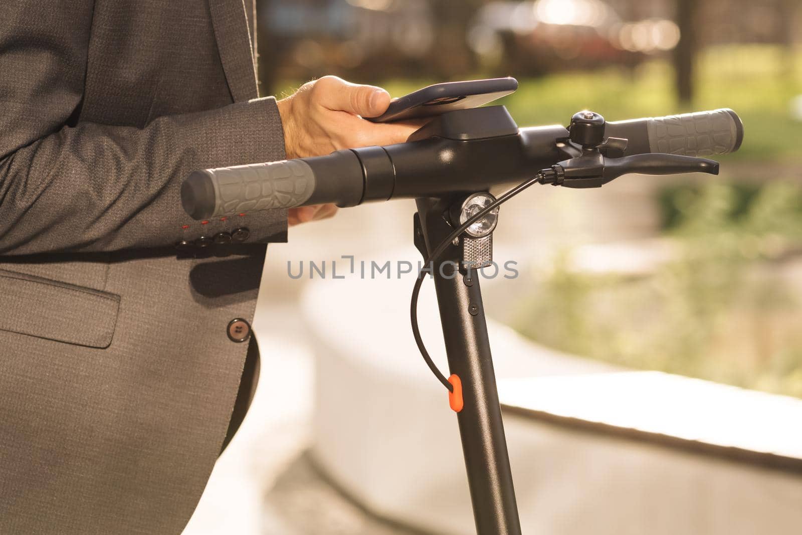 Unknown businessman in classic suit is coming up to an electric scooter and using mobile phone app. Man rents electric scooter. Eco-friendly transportation. Outdoors of business center.