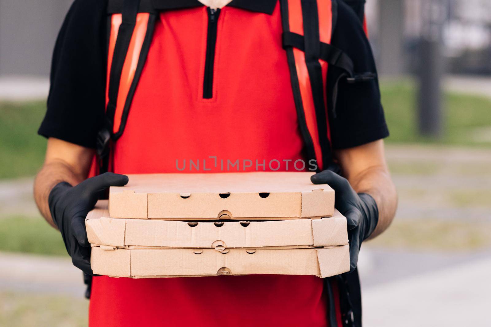 Portrait of handsome caucasian delivery man in gloves and medical mask standing at street, holding and handing carton boxes. Male courier giving pizza by uflypro