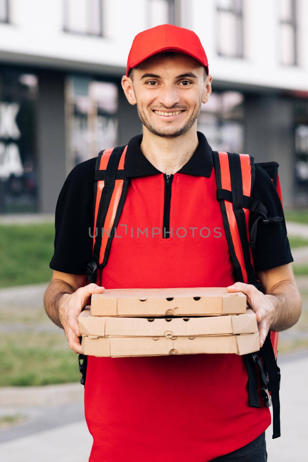 Portrait of young courier delivery man with red backpack holding pizza in carton boxes, waiting for customer in residential area. Delivery services. Excellent delivery by uflypro