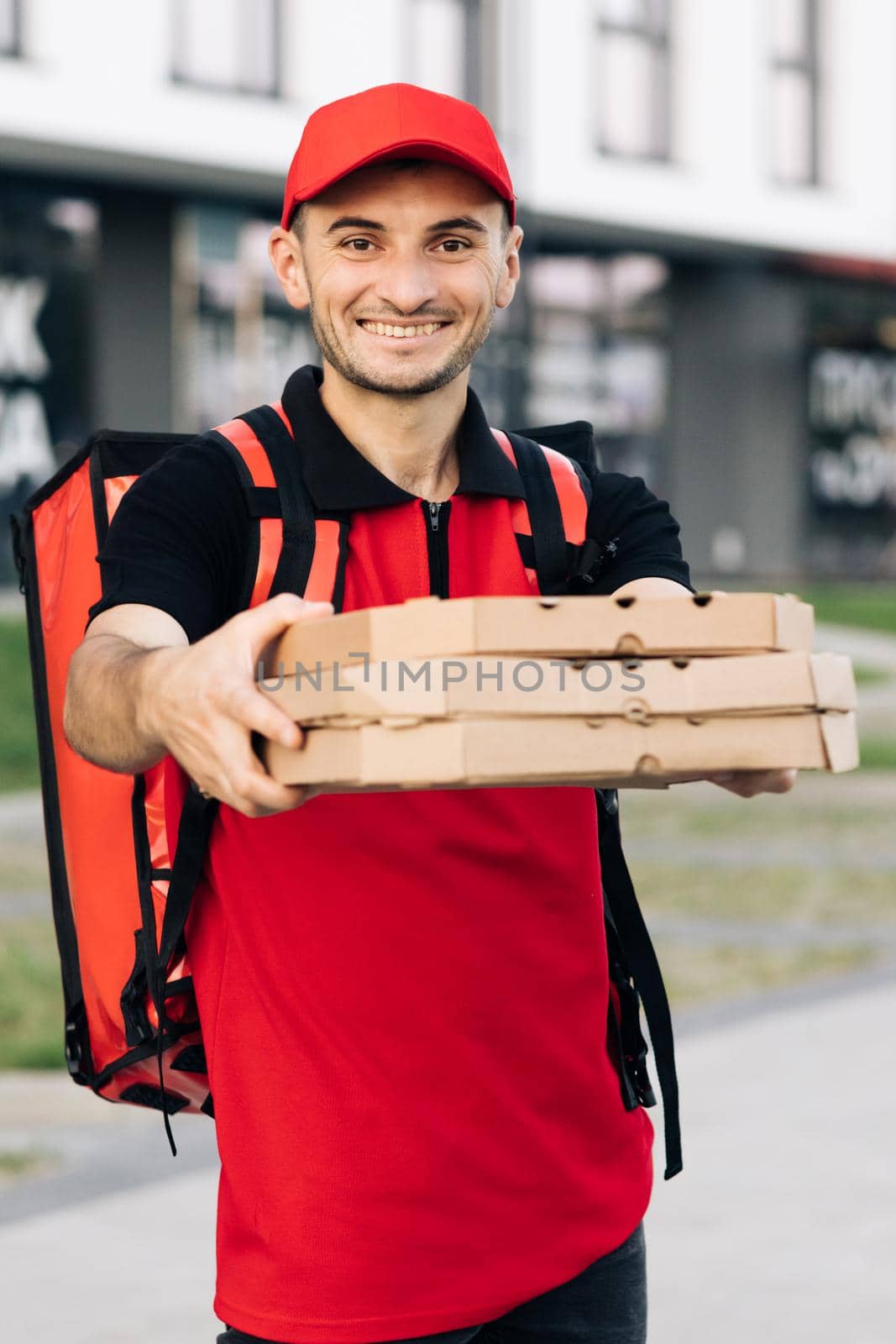 Attractive young delivery man courier in red cap smiling into camera holding pizza boxes delivering fast food around city. Outdoor portrait. Urban worker. Concept of courier, home delivery, pizza.
