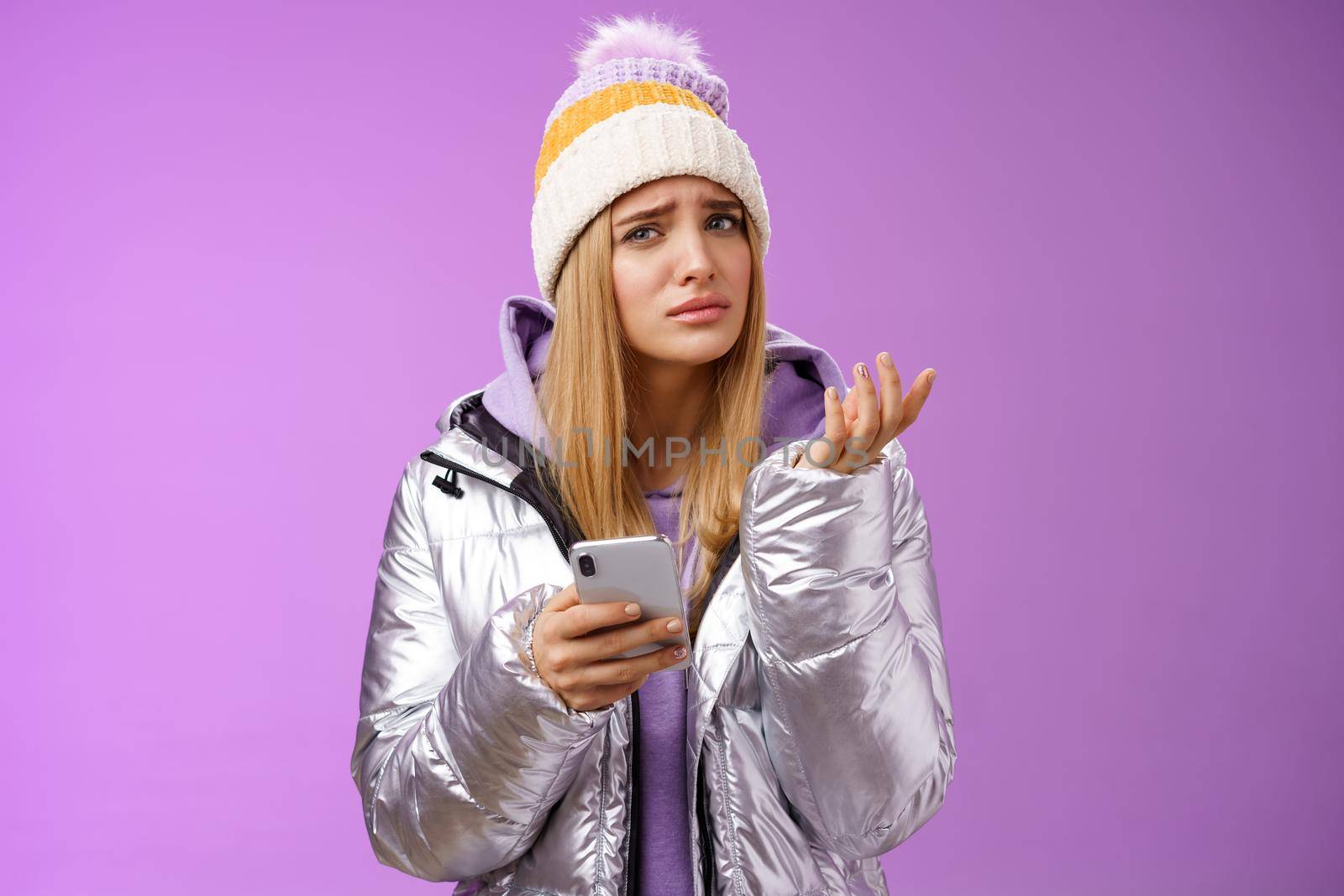 Questioned complicated cute blond girlfriend receive strange message look perplexed confused raising hand shrugging lift eyebrow cannot understand meaning holding smartphone, purple background.
