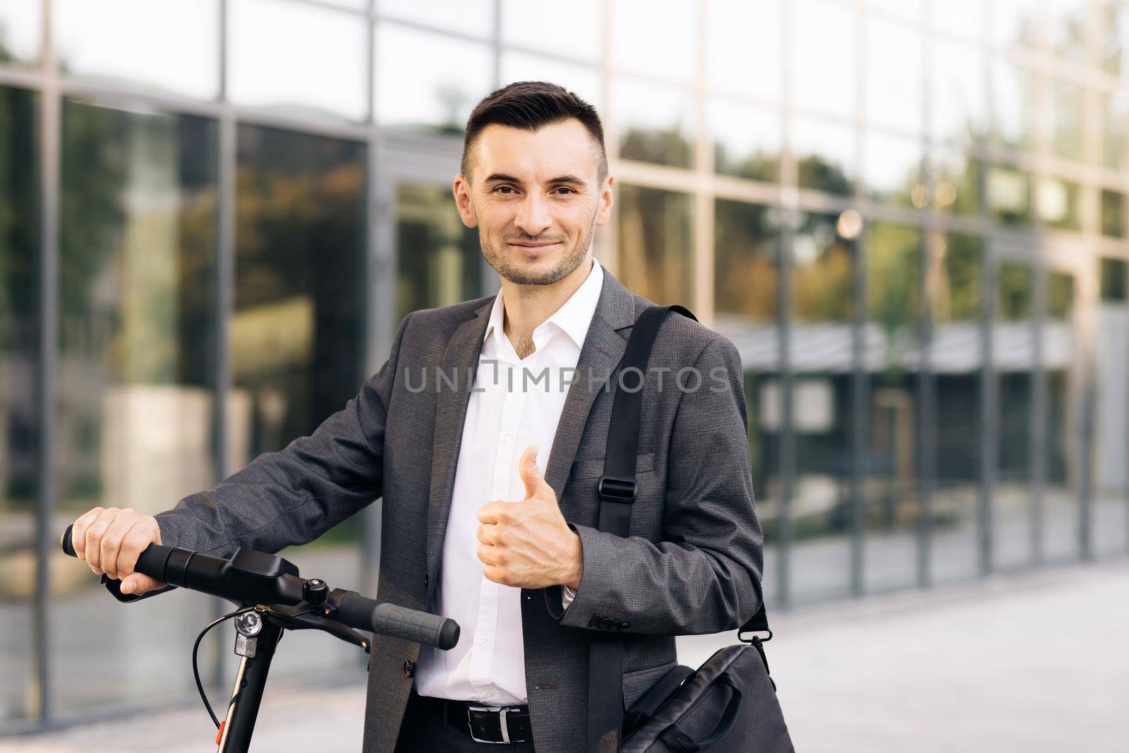 Handsome businessman with his electric scooter in front the business building. Young businessman in elegant suit standing outdoors, cheerfully smiling and showing thumb up.