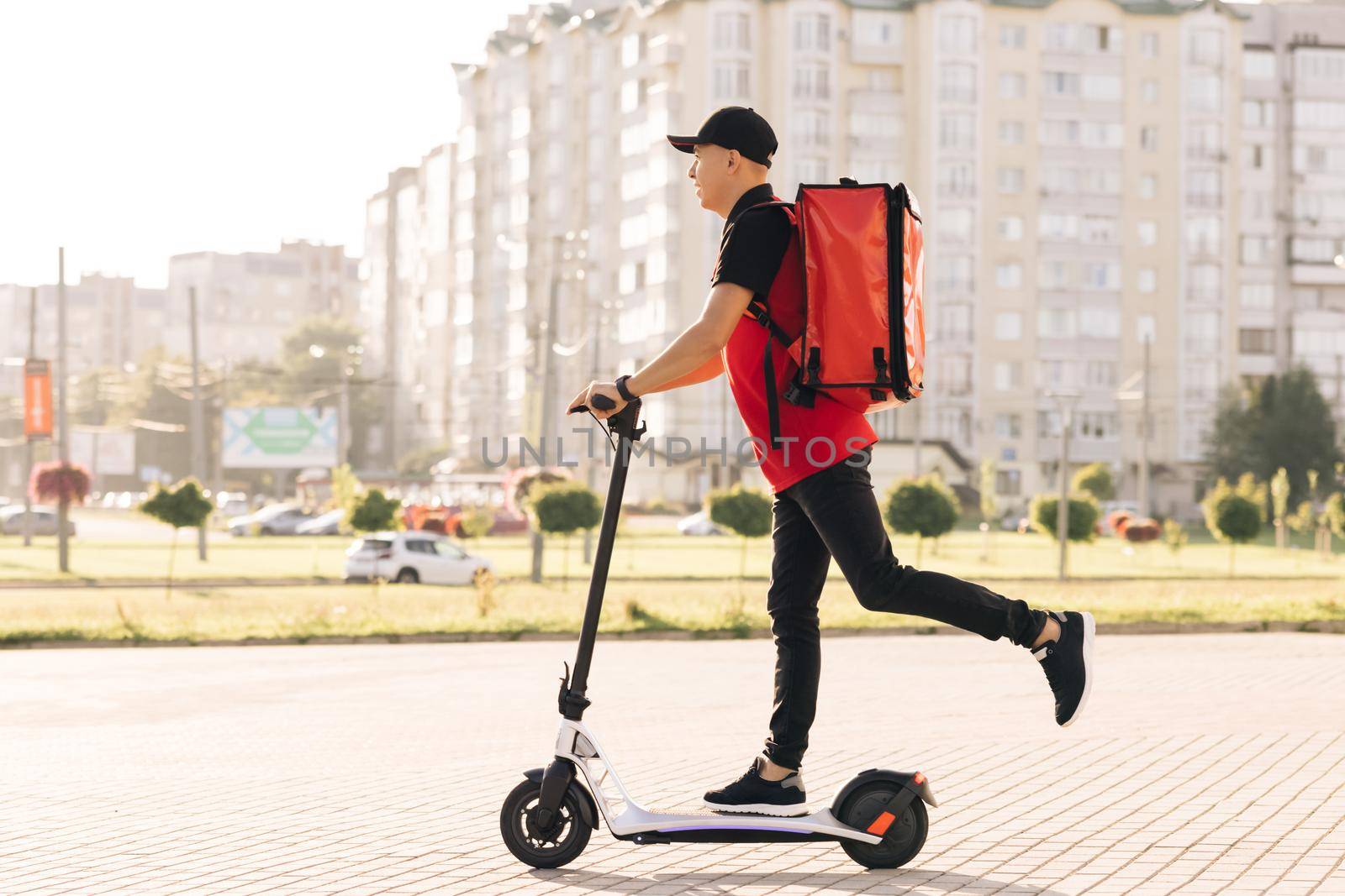 Man courier food delivery with red thermal backpack rides the street on an electric scooter deliver online order client customer. Deliveryman worker employee in red uniform. Excellent delivery by uflypro