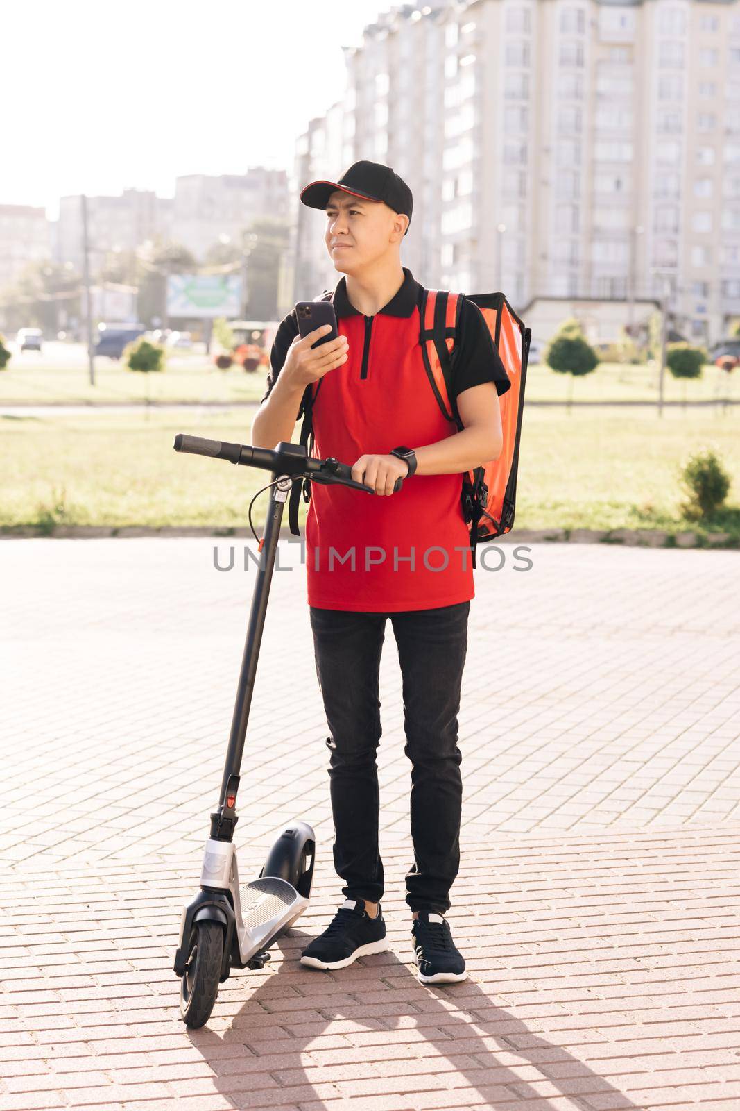 Male courier man is looking for mobile phone to deliver address of food from a restaurant. Food delivery guy navigates in phone in city with electric scooter. Eco friendly transportation by uflypro