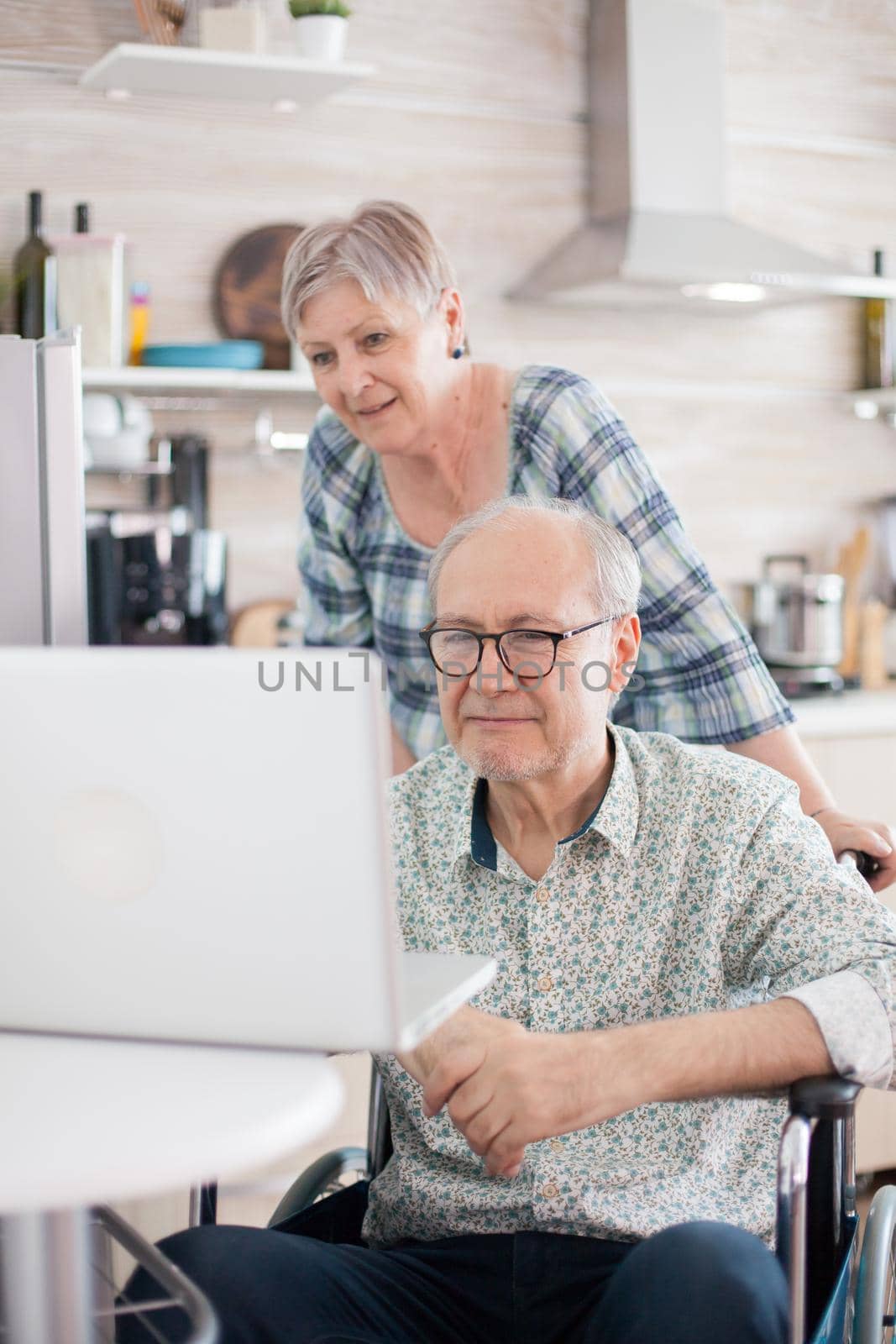 Handicapped man during video call with his family. Disabled senior man in wheelchair and his wife having a video conference on laptop in kitchen. Paralyzed old man and his wife having a online conference.