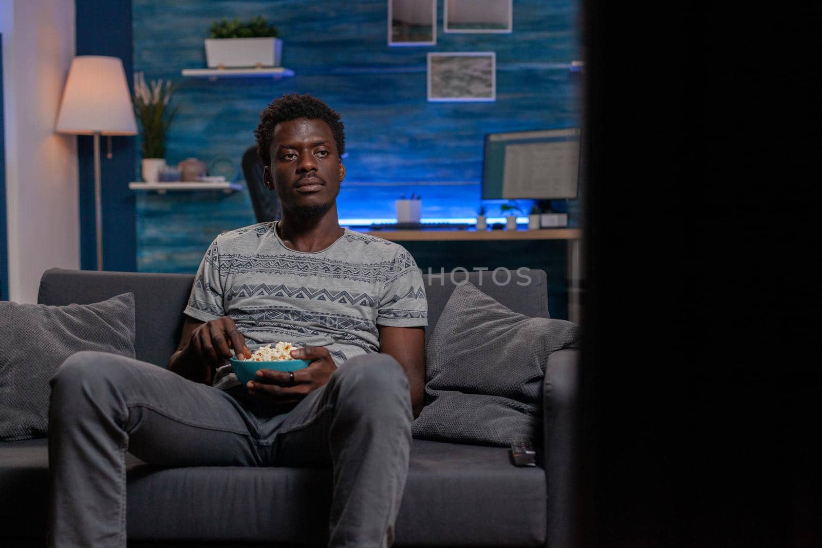 African american guy sitting on couch in living room holding popcorn bowl while watching entertainment movie on television. Young man enjoying spending free time alone in living room