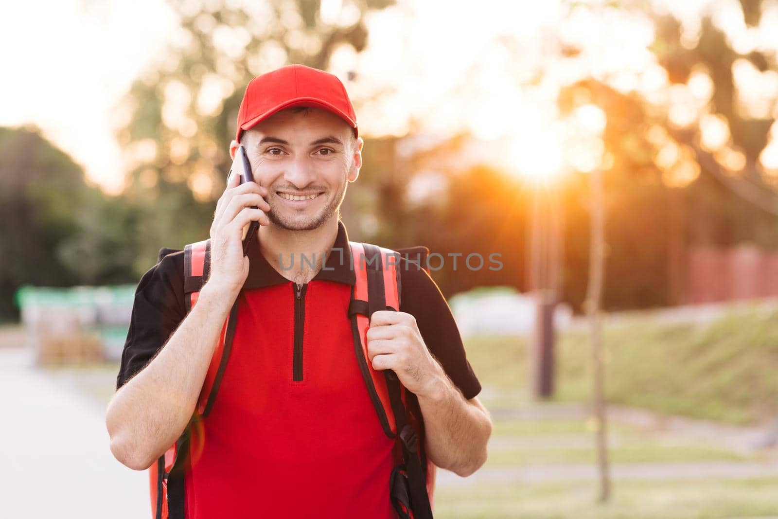Food delivery guy with red backpack deliver orders. Male courier with isothermal food case box on electric scooter arrives to the entrance to the house and calls for client by uflypro