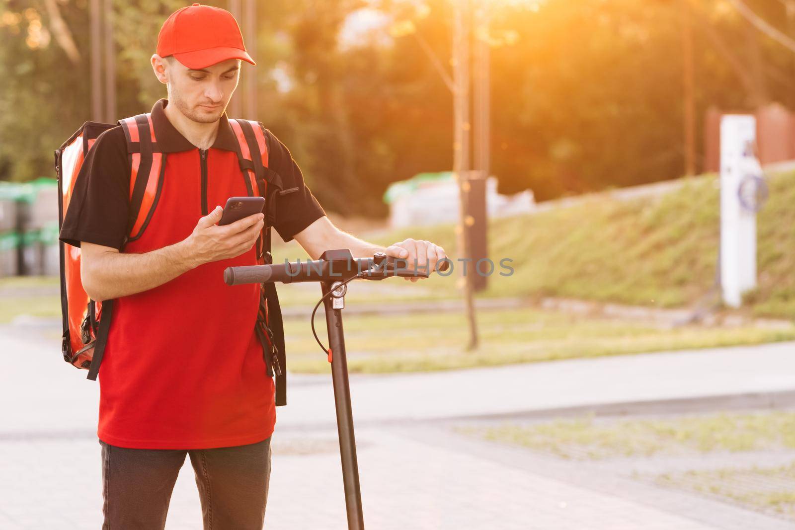 Deliveryman worker employee deliver online order client customer. Male courier food delivery with red thermal backpack looks around street with electric scooter uses smartphone navigate by uflypro