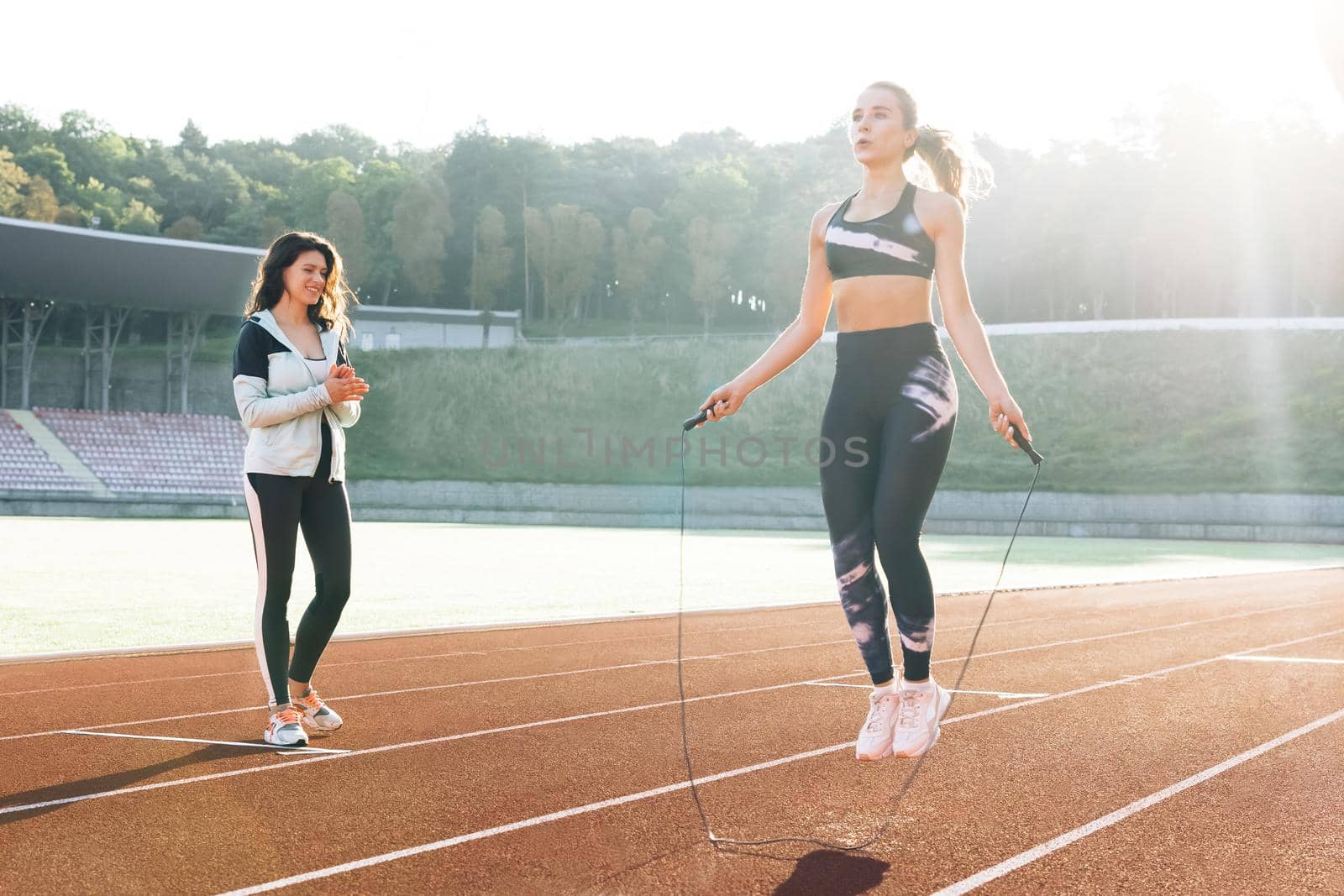 Sporty female with a good figure jumps rope on sports track of stadium. Athletic caucasian woman with personal trainer jumping rope as part of her fitness workout. Exercising strength cardio and power