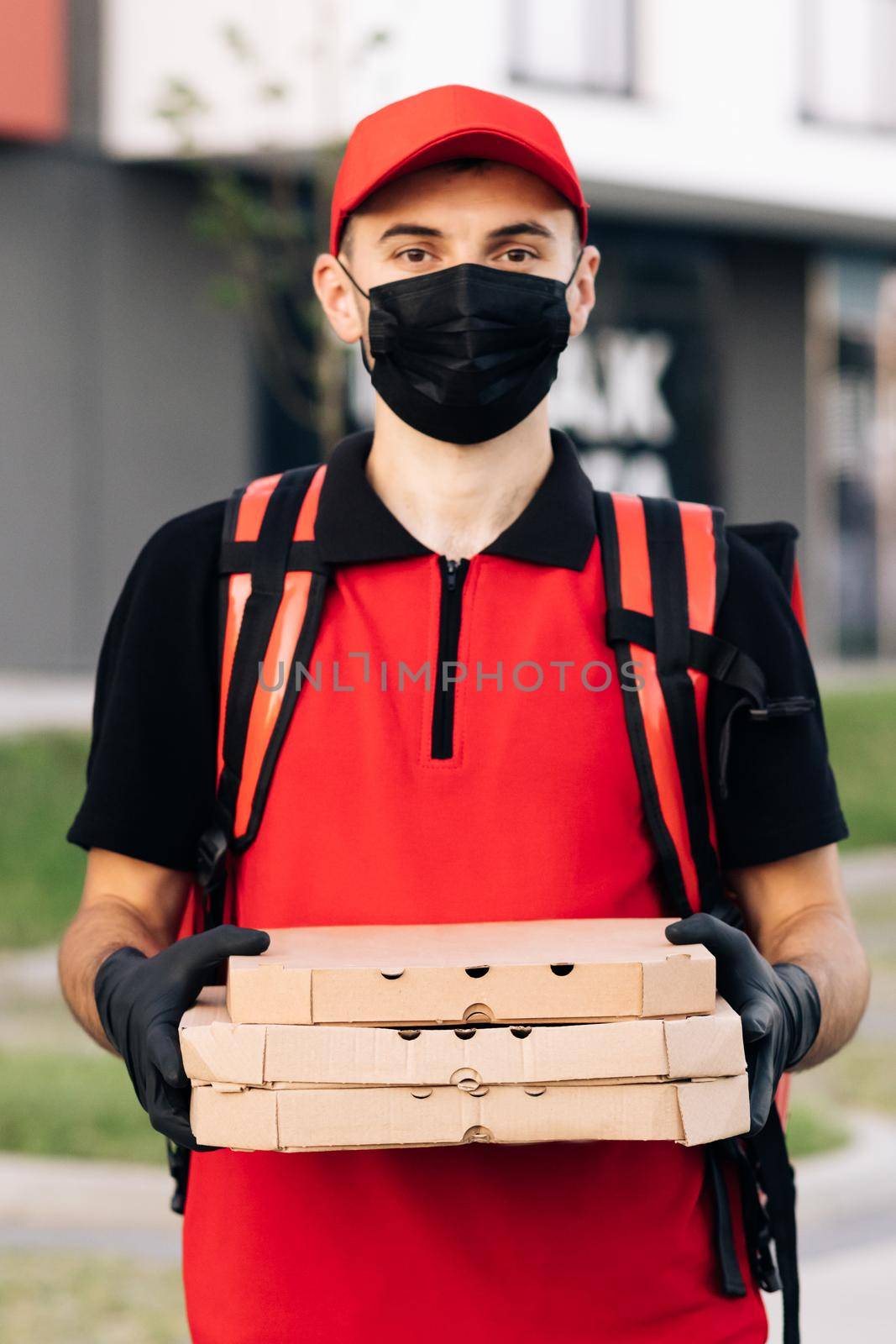 Portrait of courier delivery man with red backpack holding pizza in carton boxes, wearing protective face mask and gloves. Excellent delivery. Delivery services.