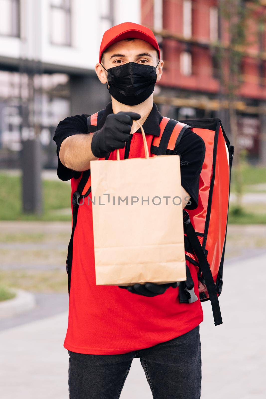 Portrait of caucasian man in red uniform, cap and medical mask from delivering service turning and smiling to camera with packet of food. Male courier with eco grocery packet by uflypro