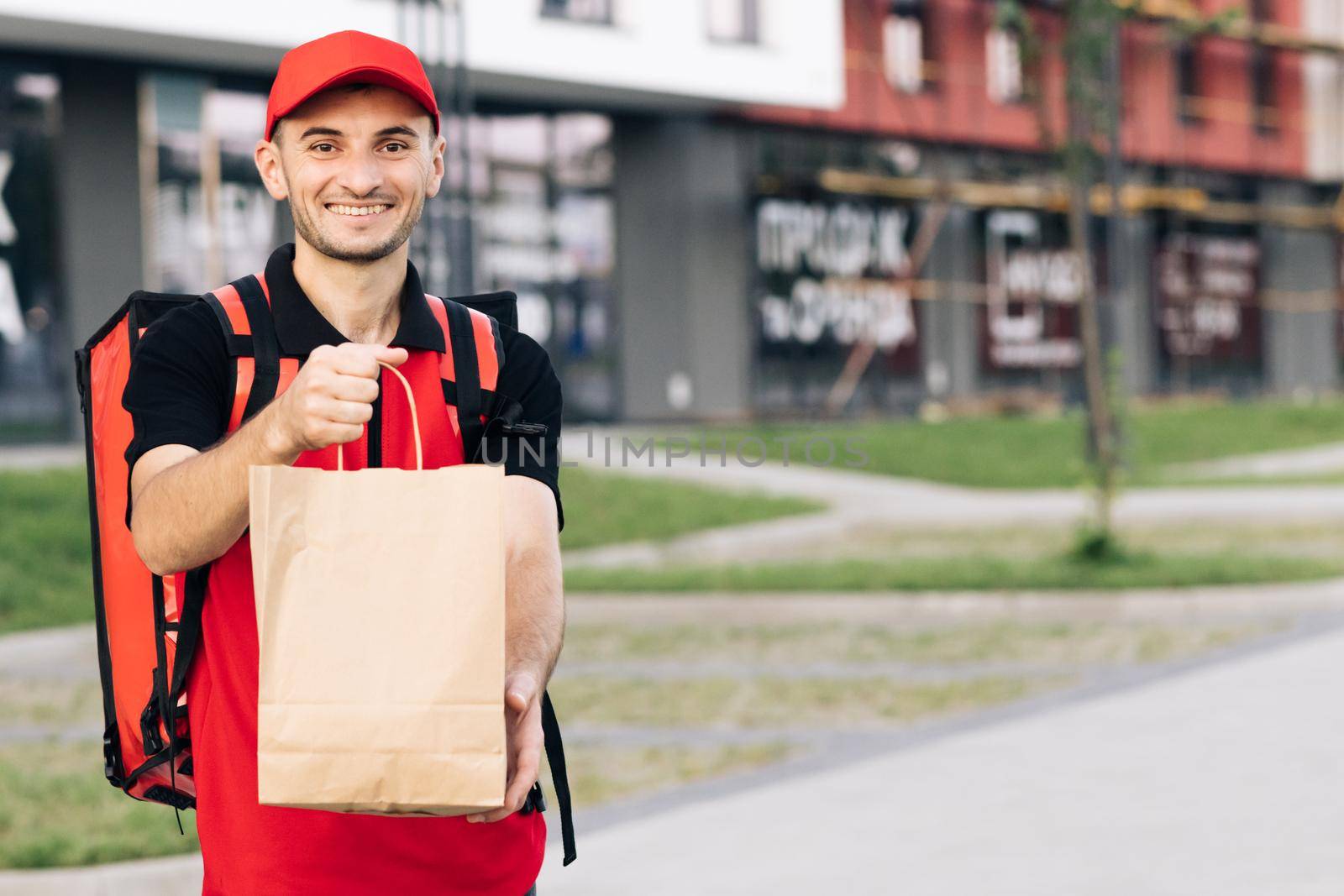 Happy young courier is proud of his job smiling standing in the street. Home delivery. Outdoor portrait of delivery man with red uniform holding food bags waiting for customer.