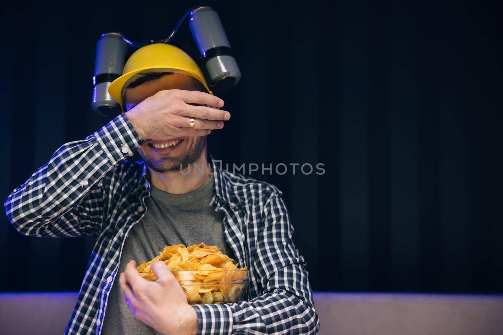 Caucasian man rests at home and watches TV shows or sports news on TV screen. Man with beer helmet on the head eats chips while sitting at home on couch, in evening he watches movies at home on TV.