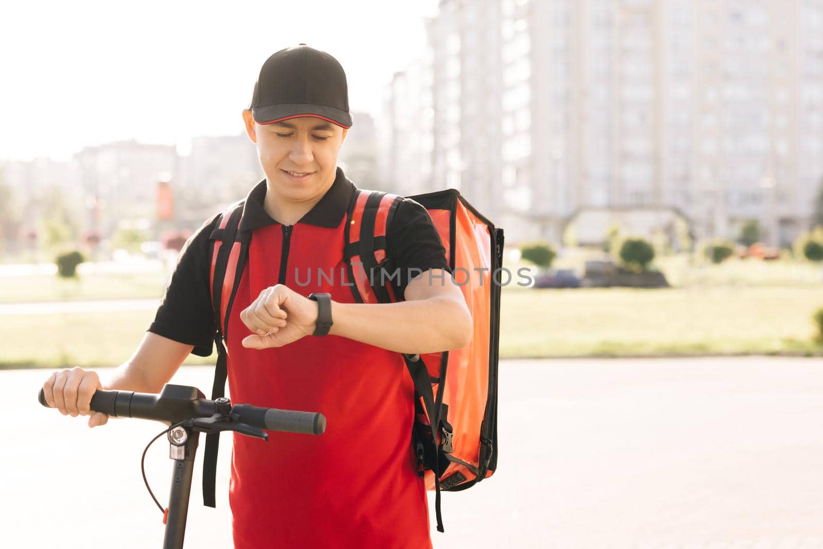 Positive deliveryman worker employee deliver online order client customer. Smiling man courier food delivery with red thermal backpack walks street with electric scooter.