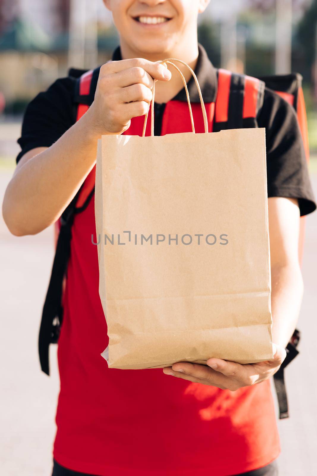 Happy delivery worker holding packet with food looking at the camera and smiling. Close up portrait of positive young man courier person. Delivery service door to door.