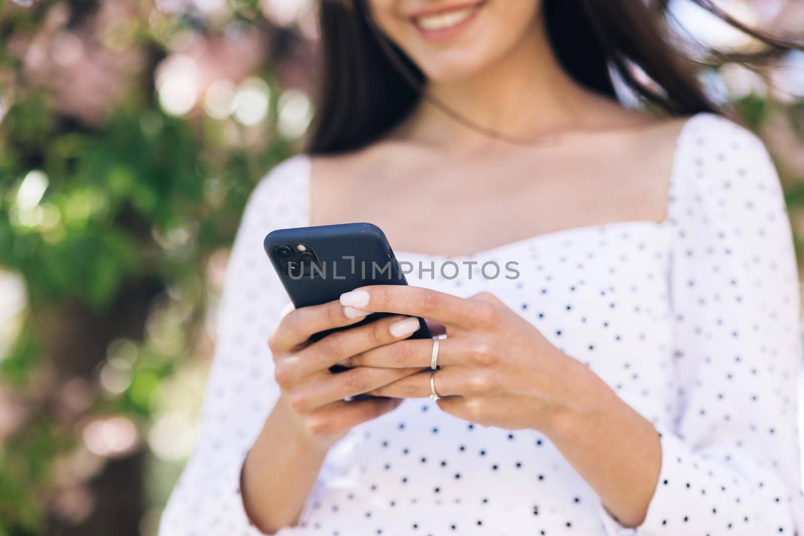 Happy hipster woman typing by mobile phone outdoors. Cheerful girl with smartphone in park on a background of sakura trees. Smiling lady holding cellphone in hands outside by uflypro