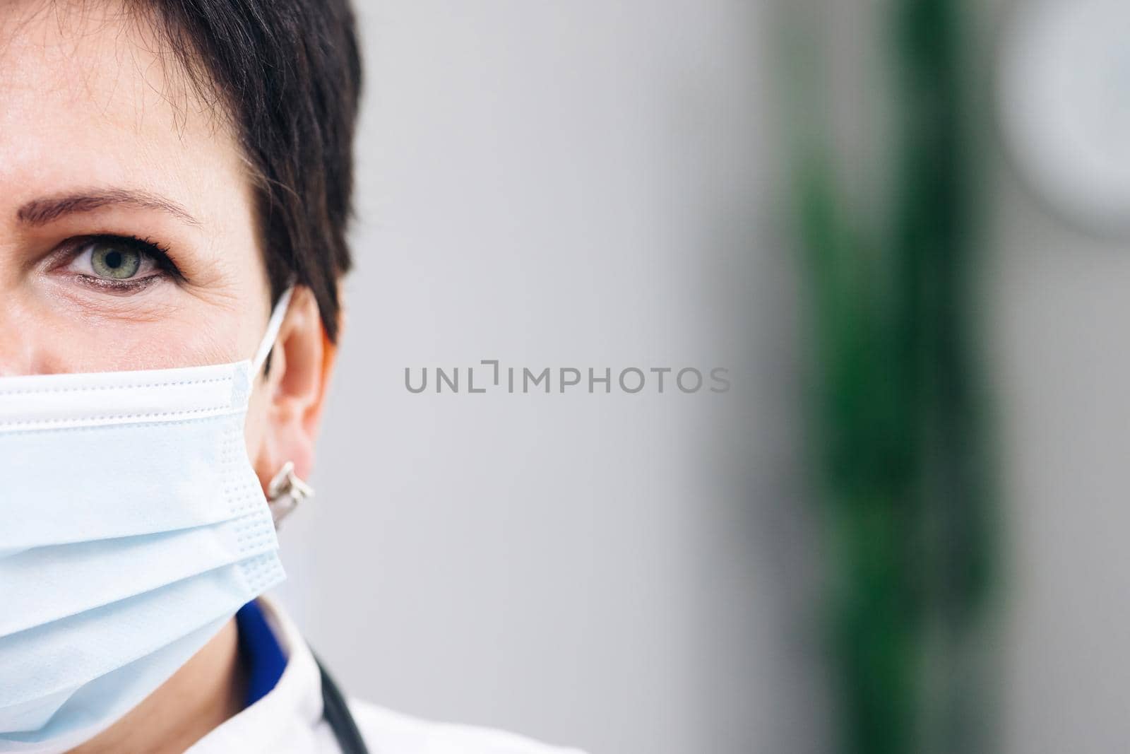 Close up half face portrait of Senior Family Medical Doctor in Face Mask. Successful Physician in White Lab Coat Looks at the Camera in Hospital Office. Covid-19 Pandemic Concept of Infectious Virus