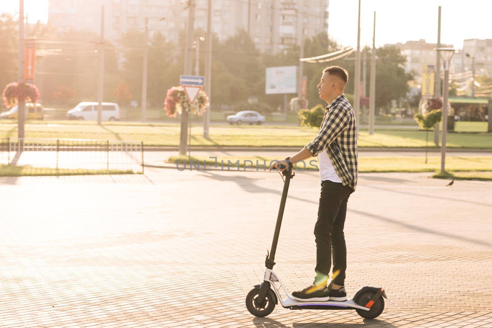 Young smart hipster man riding a fast modern electric scooter near huge contemporary construction in open landscape on country road. Eco transportation. Fast speed driving electric transport by uflypro