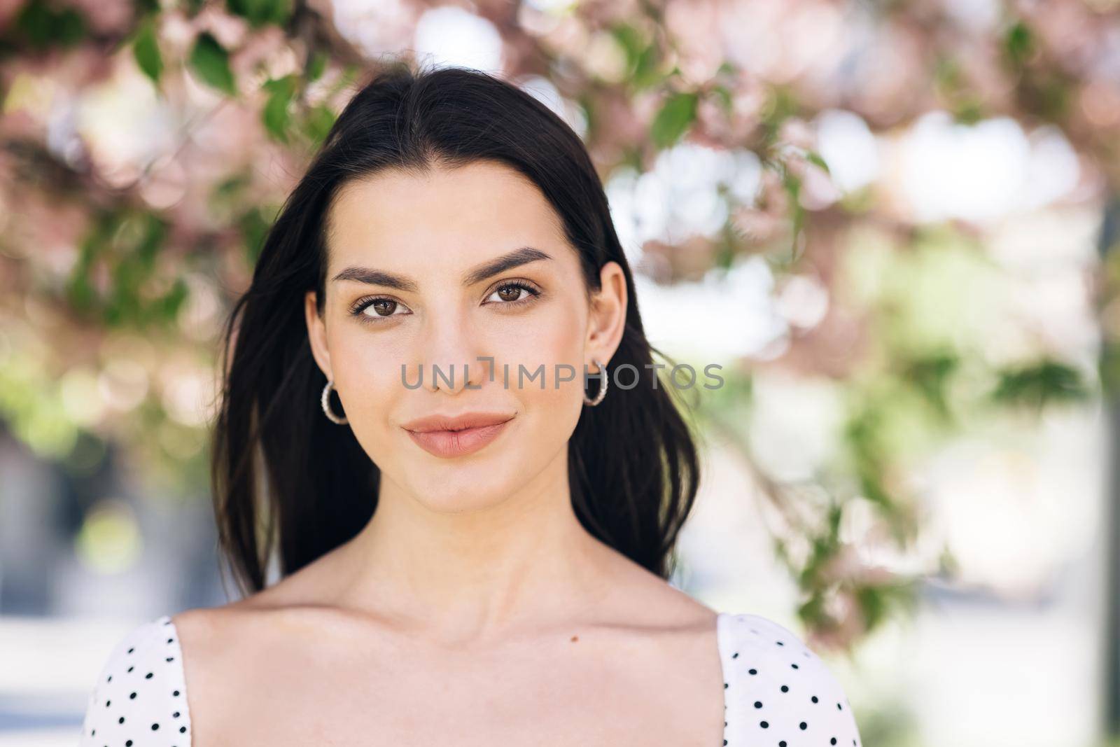 Perfect model with creative vivid makeup and pink lipstick on lips and hairstyle posing outside happy looking at camera. Spring blossom of sakura tree in city park by uflypro