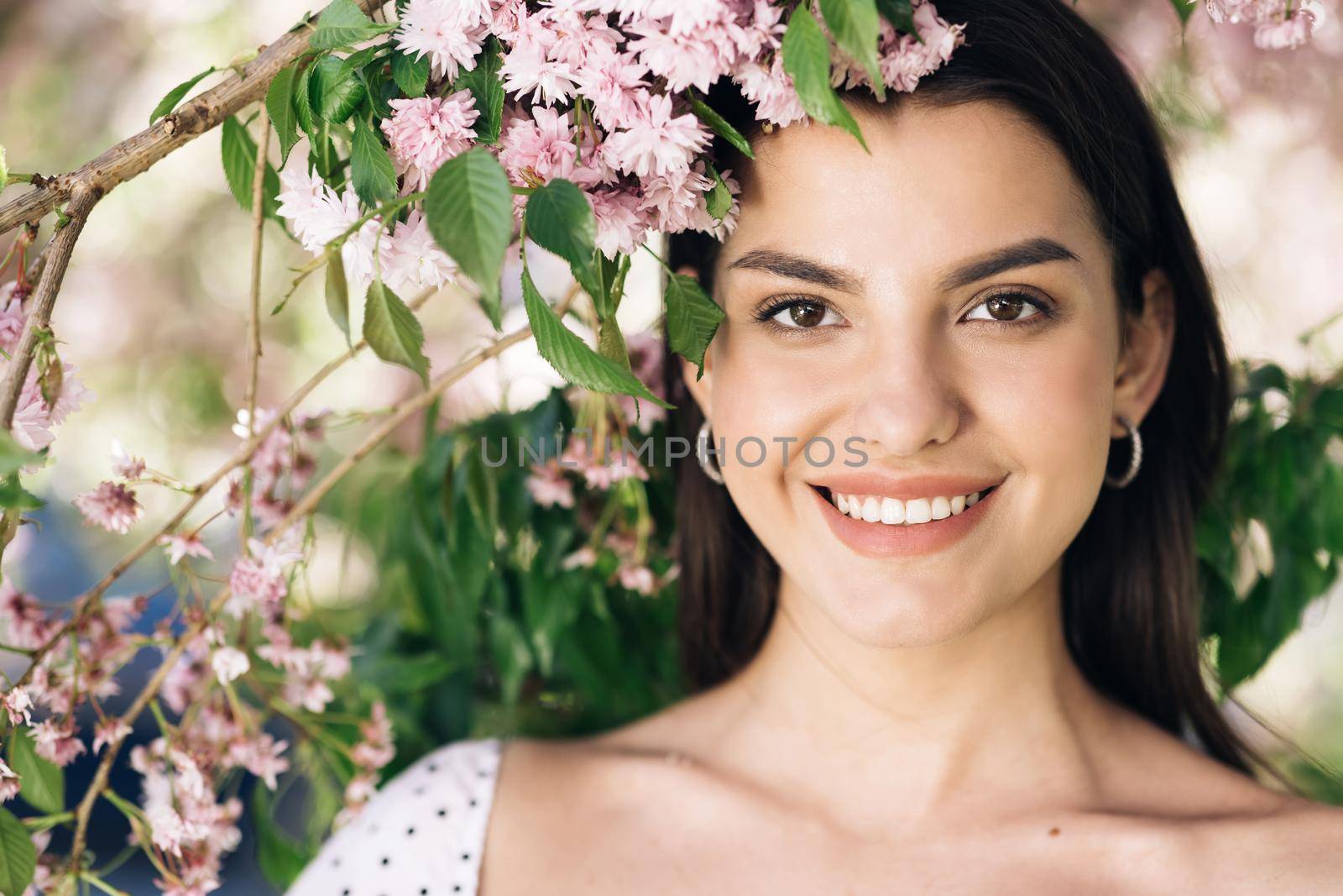 Perfect model with creative vivid makeup and pink lipstick on lips and hairstyle posing outside happy looking at camera. Spring blossom of sakura tree in city park.