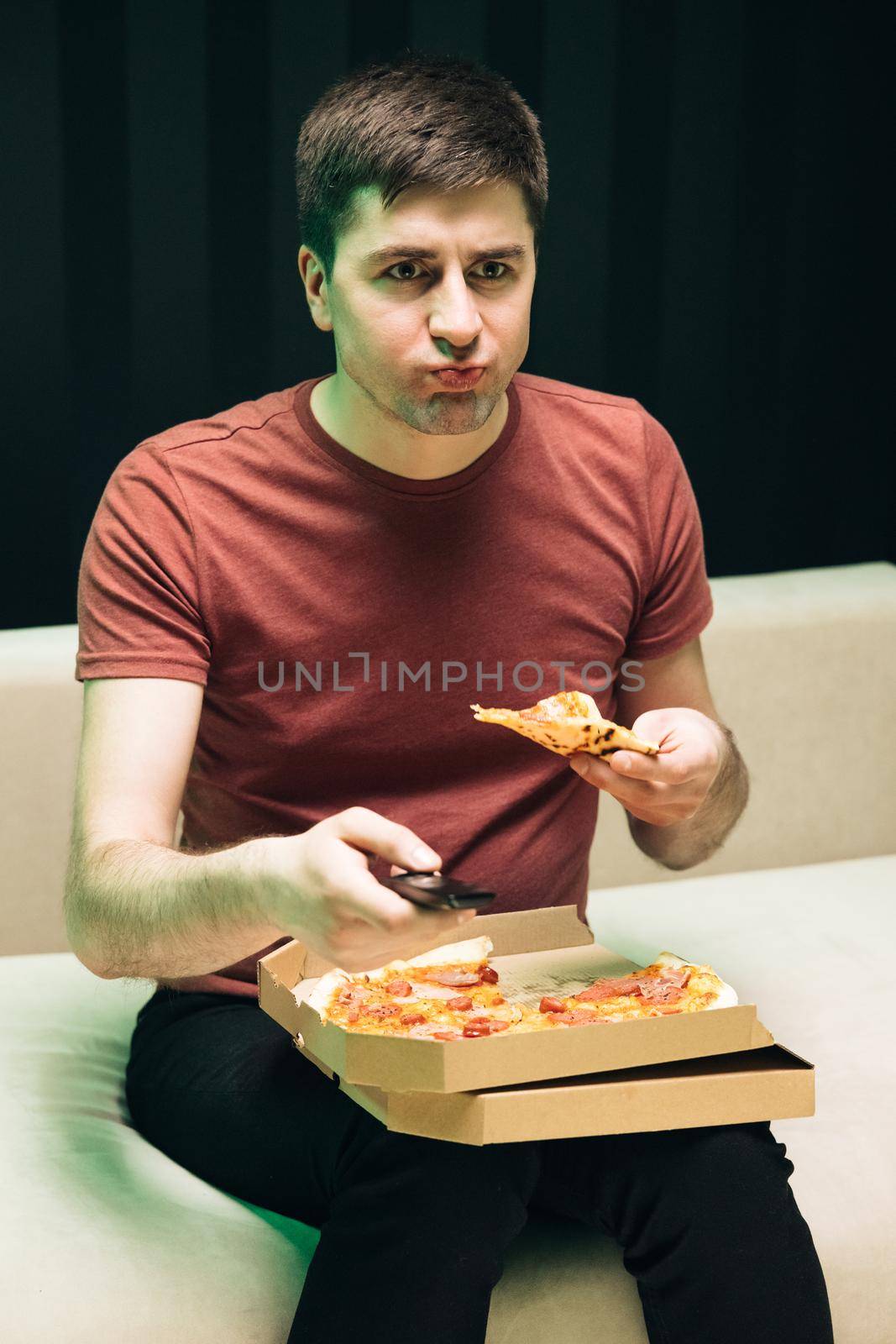 Young hipster man eating pizza watching favorite comedy on tv while relaxing at sofa in the living room. Happy guy eating Italian food